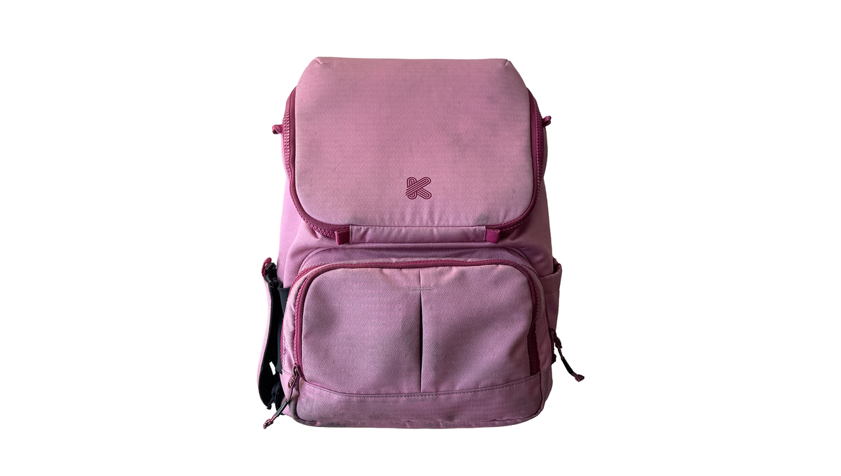 A dusty pink backpack with a front flap pocket and a lower zippered compartment, featuring a small embroidered pink 'K' logo on the front, set against a plain white background.