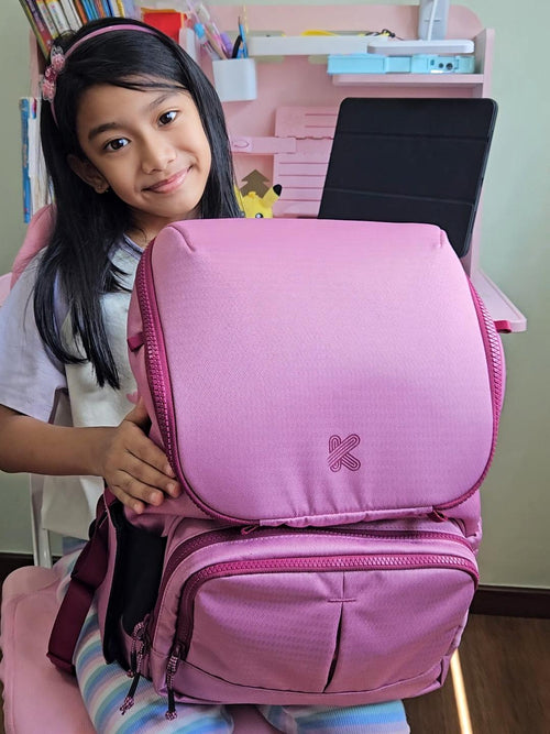 Young girl holding a pink backpack with a visible brand logo in a classroom setting.