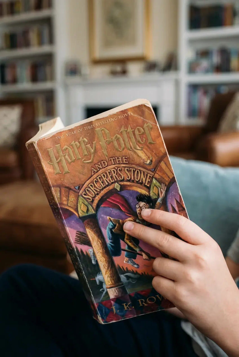 A person holding a copy of Harry Potter and the Sorcerer's Stone in a cozy living room setting with bookshelves in the background.