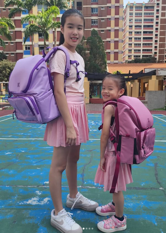 Two children with backpacks standing on a playground with buildings in the background