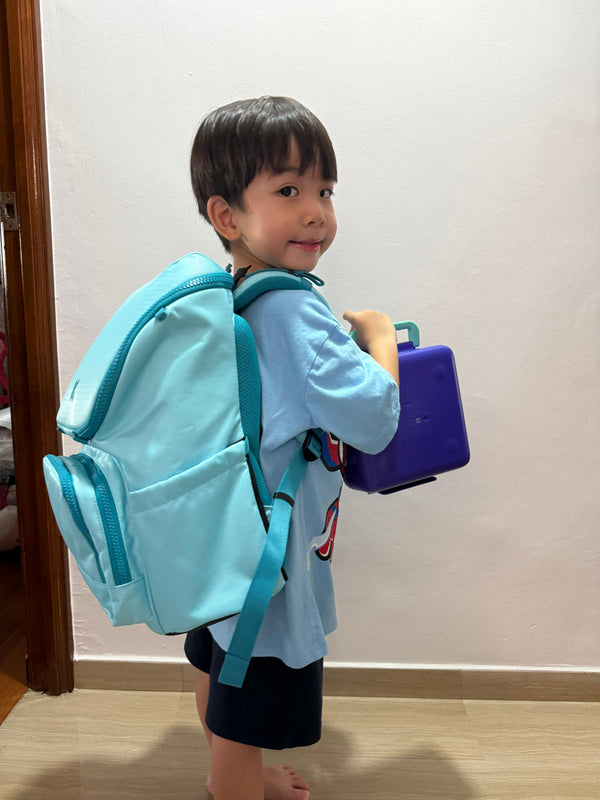 Child with a light blue backpack and purple book standing indoors.