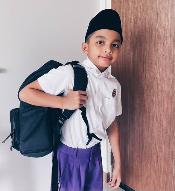 Child in school uniform with a backpack standing in front of a wooden door.
