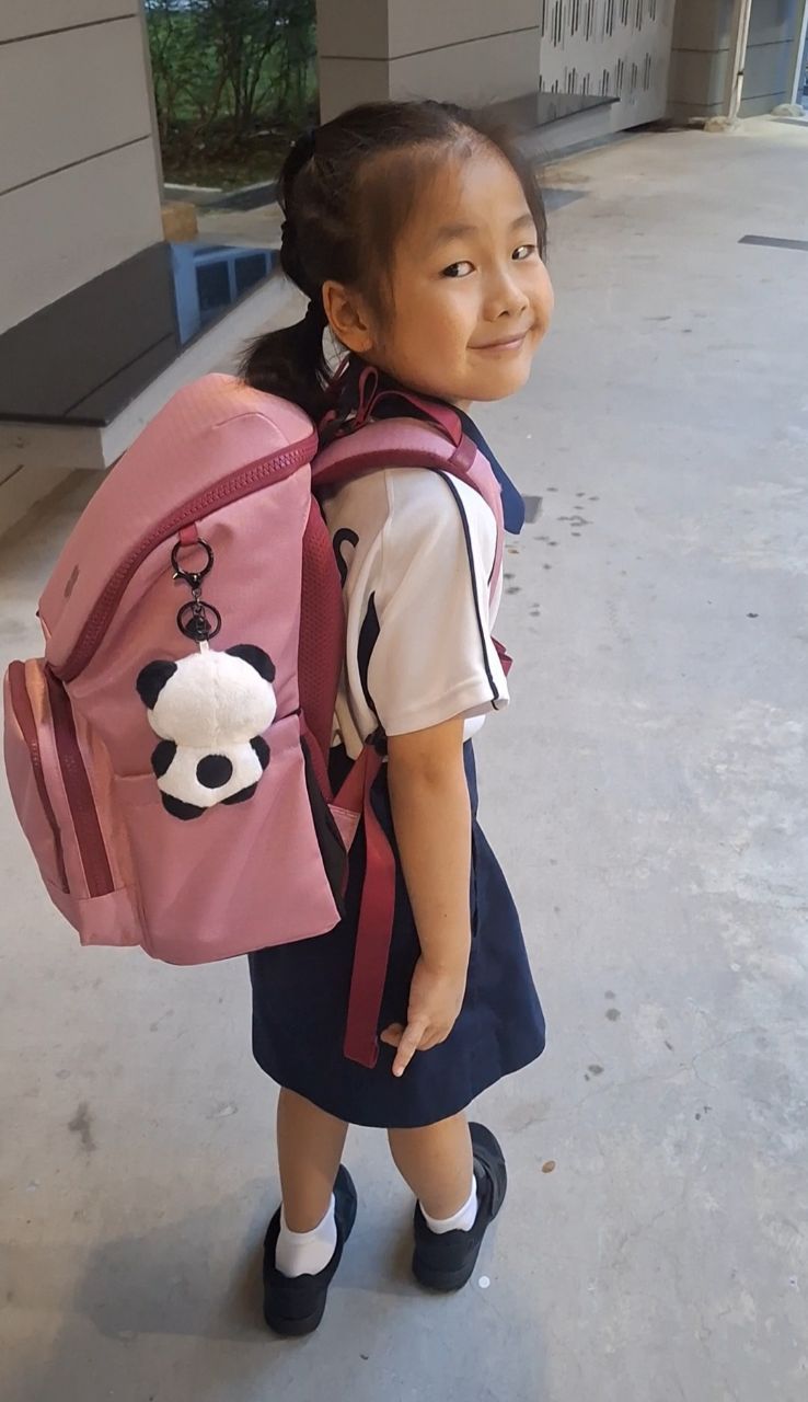 Young girl with a pink backpack featuring a panda charm, standing on a concrete surface.