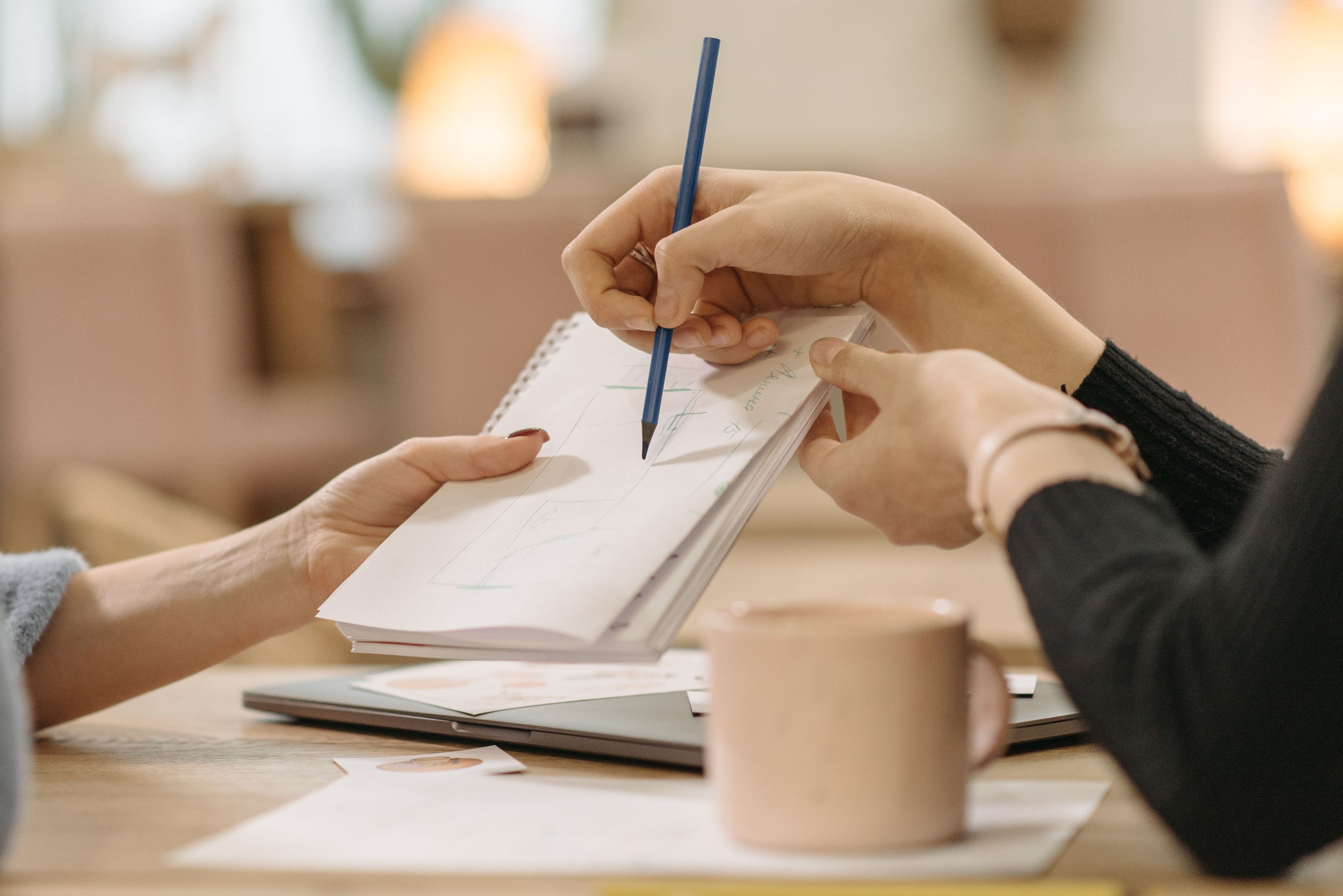 A close-up lifestyle shot of two people collaborating at a table, with one person holding a pen and writing in a notebook while the other holds it open. A pink mug sits on the table in the foreground, and the background is a softly blurred indoor setting.