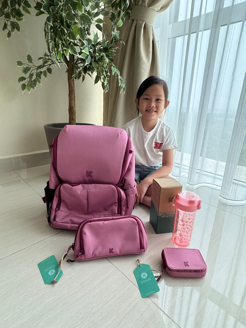 Child sitting on the floor with pink backpack and accessories in a bright room.