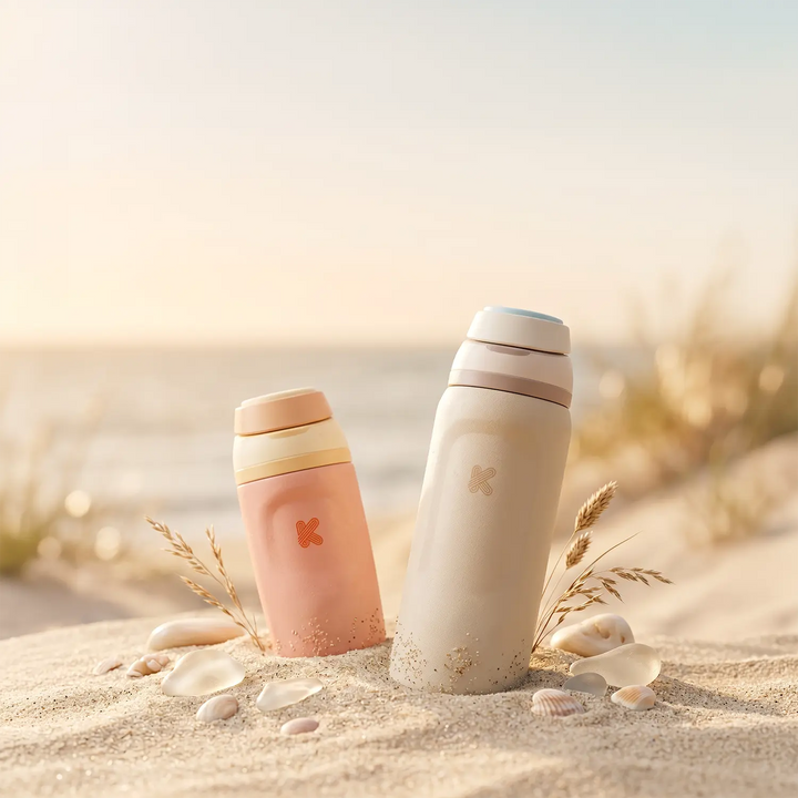 Two insulated water bottles standing in sand on a beach, with shells and dried grass around them. One bottle is pastel peach with a cream lid, and the other is off-white with a pale pink lid.