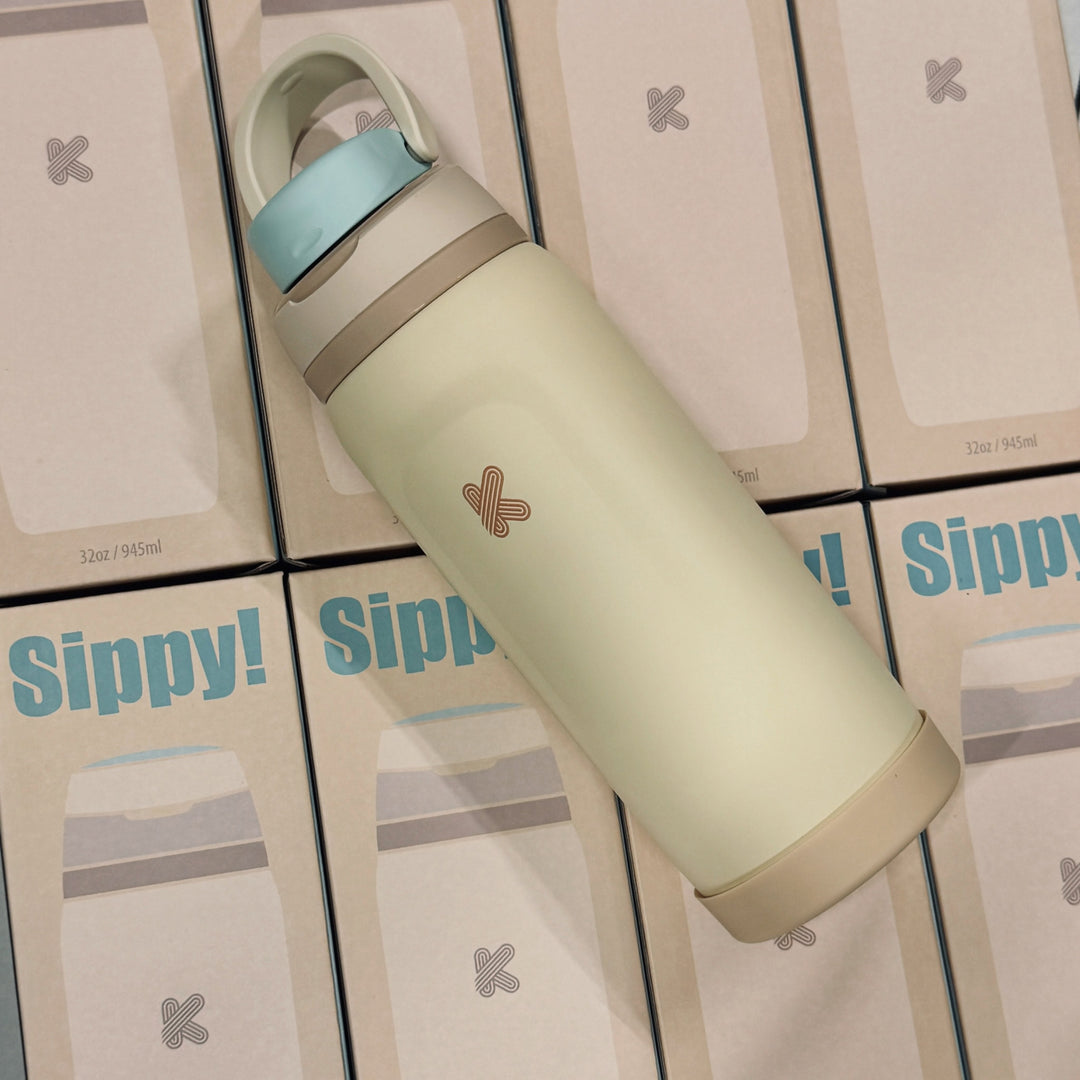 A reusable insulated bottle with a pale yellow body, light blue lid, and beige accents, placed diagonally on top of multiple Sippy! product boxes.