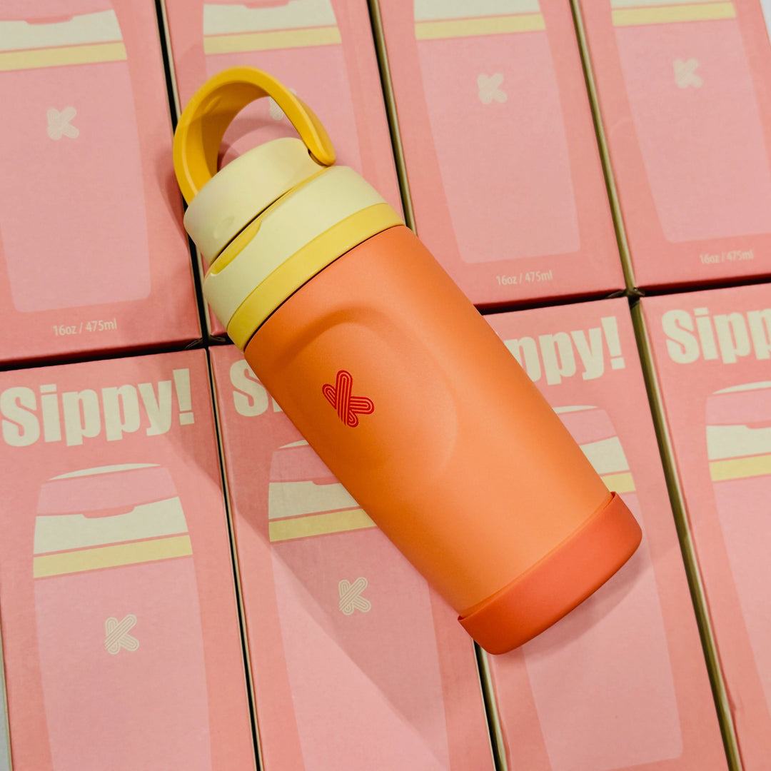 An orange insulated water bottle with a yellow cap and handle, resting diagonally on a stack of pink Sippy! product boxes.