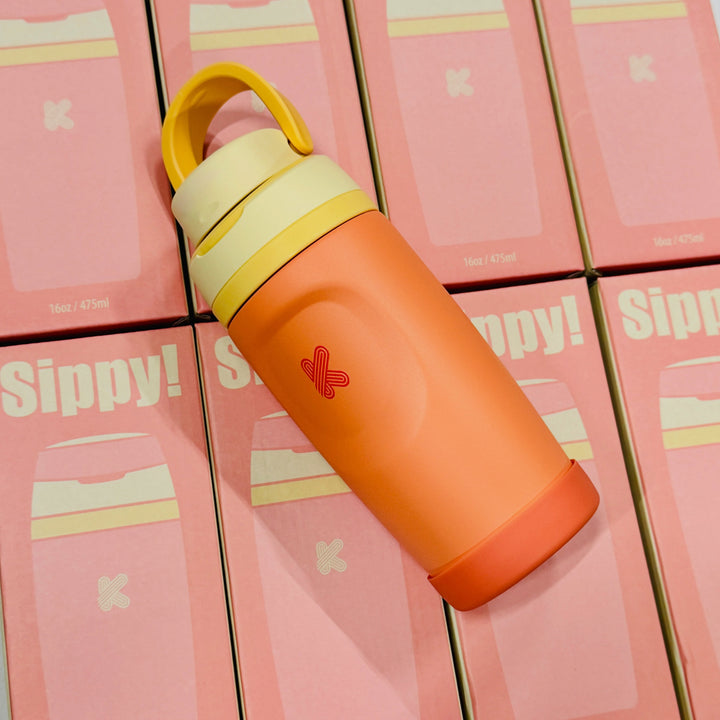 An orange insulated water bottle with a yellow cap and handle, resting diagonally on a stack of pink Sippy! product boxes.