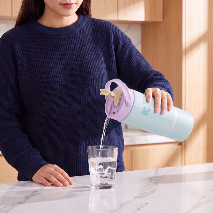 A woman in a navy blue knit sweater pours water from a pastel light blue and lavender insulated water bottle with a butterfly design into a clear glass on a kitchen counter.