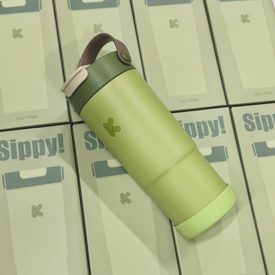 A light green insulated travel tumbler with a dark green lid and a brown carrying loop, placed diagonally on top of several stacked Sippy! product boxes.
