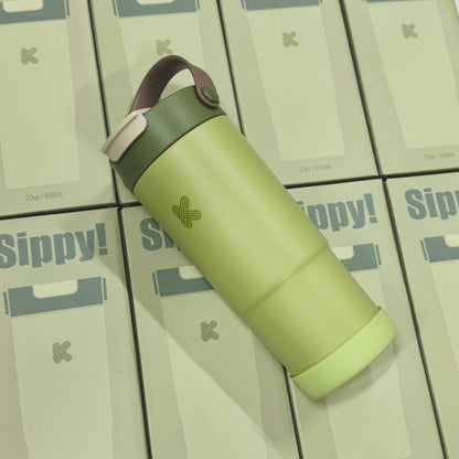A light green insulated travel tumbler with a dark green lid and a brown carrying loop, placed diagonally on top of several stacked Sippy! product boxes.