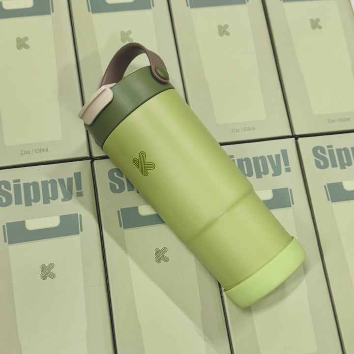 A light green insulated travel tumbler with a dark green lid and a brown carrying loop, placed diagonally on top of several stacked Sippy! product boxes.