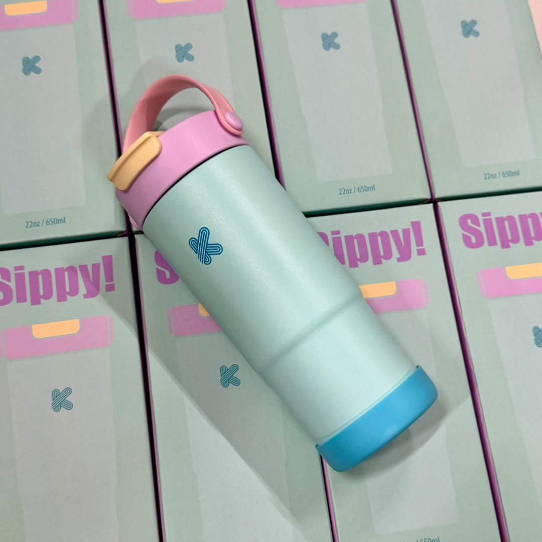 A reusable insulated bottle with a pale mint body, pink lid and handle, and a blue base, placed diagonally on top of the packaging boxes.